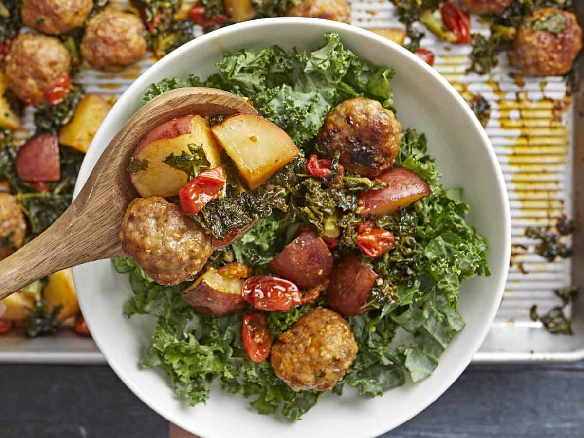 Overhead shot of Sausage Meatball and Kale Sheet Pan Meal in a white bowl with a wood spoon serving a portion into the bowl.