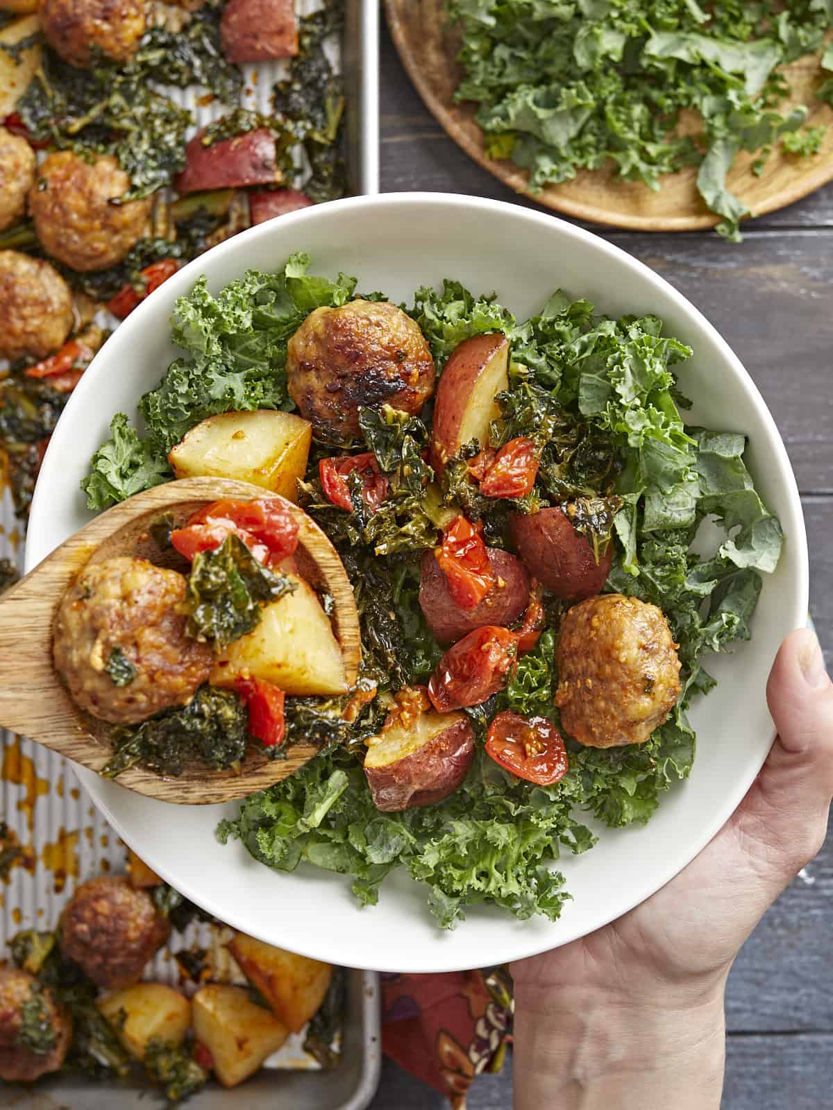 Overhead shot of Sausage Meatball and Kale Sheet Pan Meal in a white bowl with a hand holding the bowl and a wooden spoon serving a portion into the bowl.