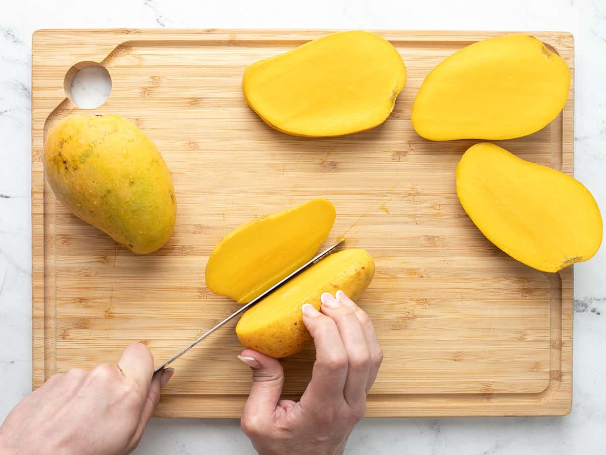 Overhead shot of one hand holding a mango while the other hand slices it on a wooden cutting board. There are other mango slices on the board.