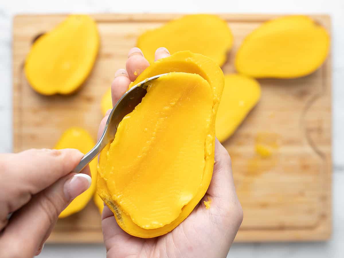 Hand holding a mango half and scooping out the flesh with a spoon.