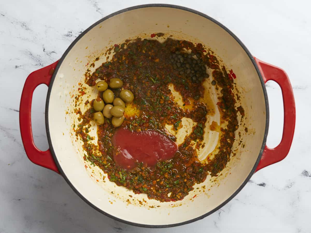 Overhead shot of sofrito, tomato sauce, olives and capers in a red Dutch oven.