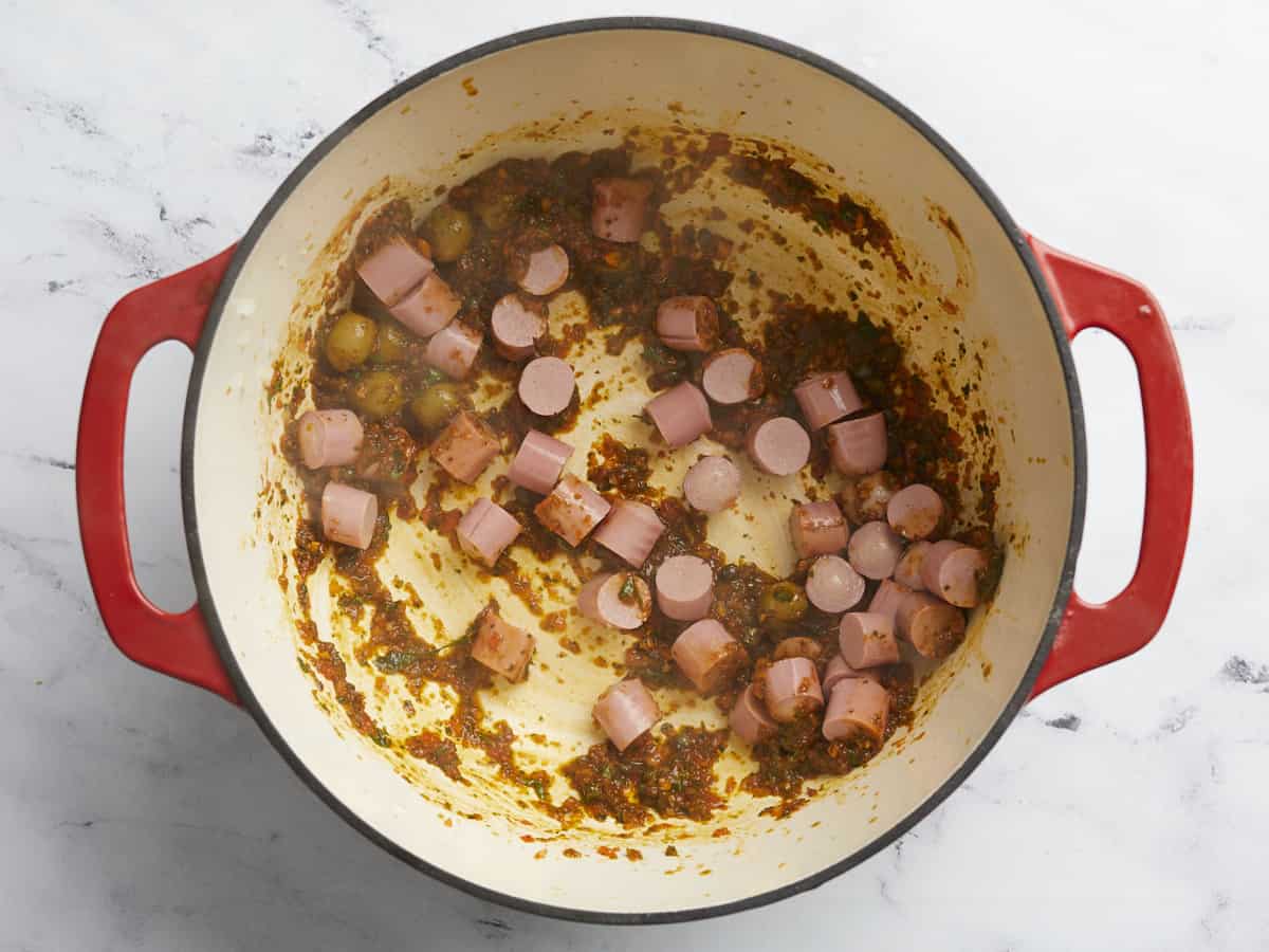 Overhead shot of salchichas added to sofrito, tomato sauce, olives and capers in a red Dutch ov