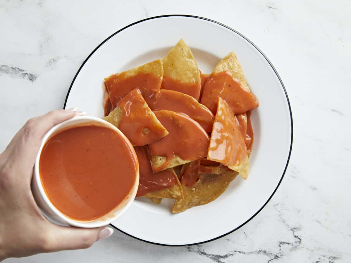 Overhead shot of fried tortillas served on a white plate with red sauce being drizzled on top.