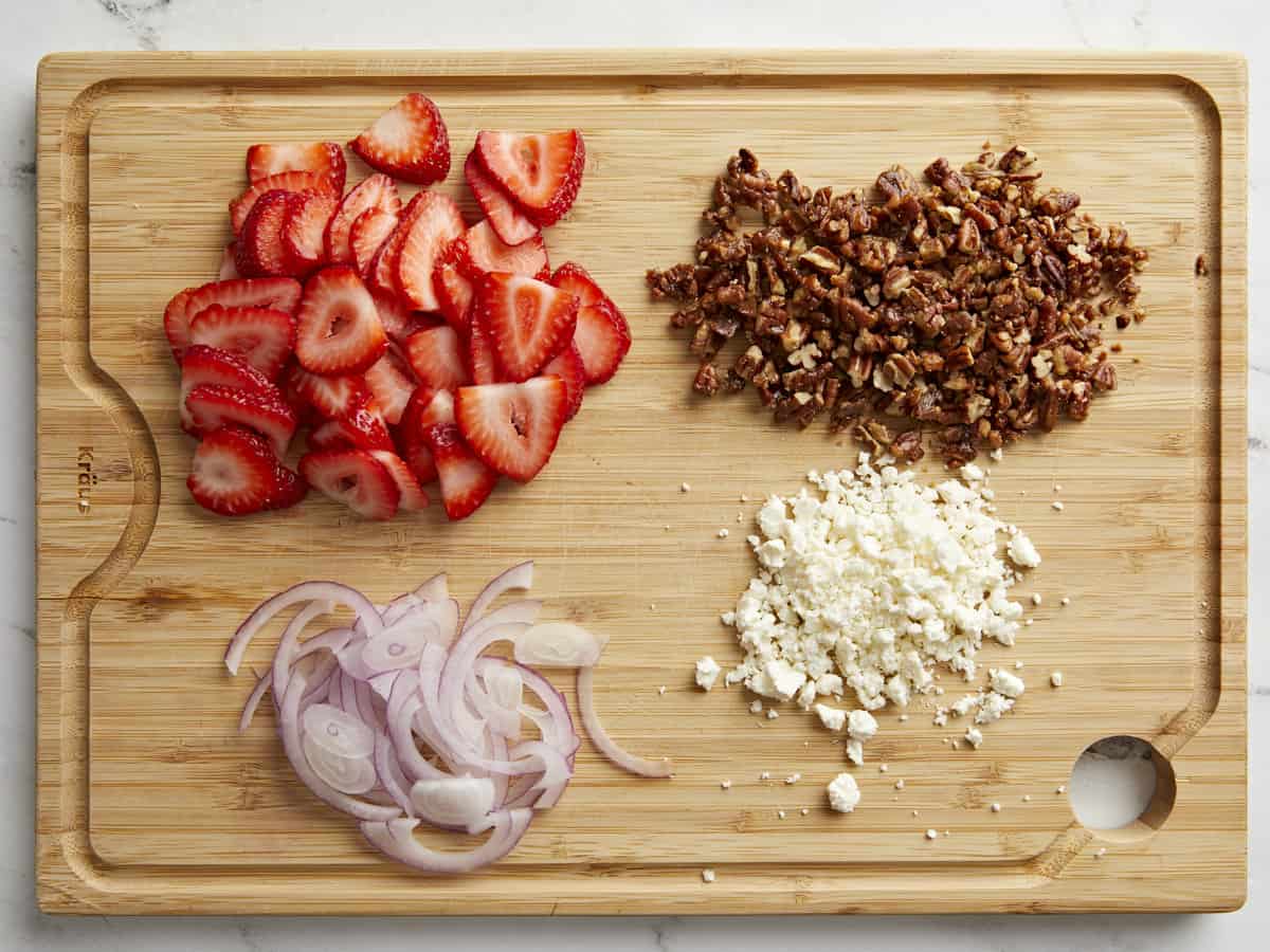Sliced strawberries, sliced red onion, feta cheese, and canided pecans on a wood cutting board