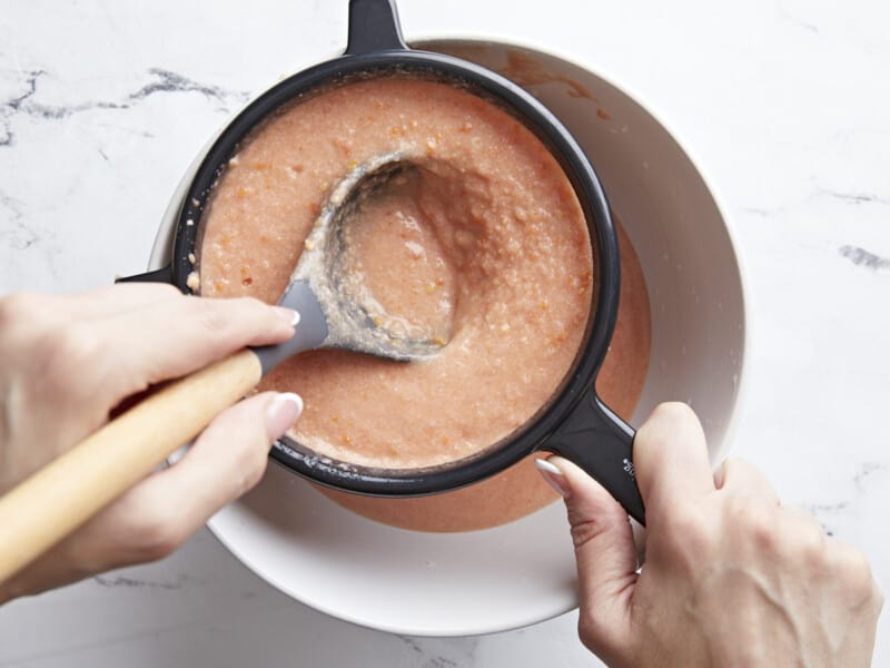 Overhead shot of a hand using a gray ladle to push Salmorejo through a fine mesh sieve with a black handle into a white bowl.