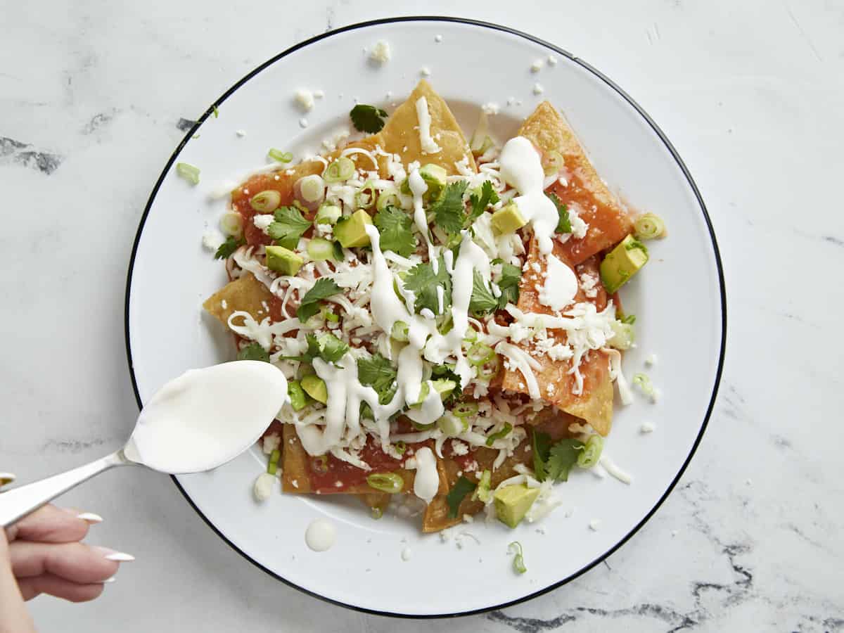 Overhead shot of chilaquiles served on a white plate with sour cream being drizzled on top.