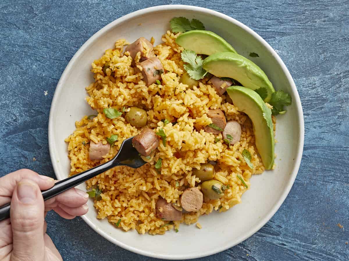 Overhead shot of Arroz Con Salchichas on a plate with a hand holding a forkful.