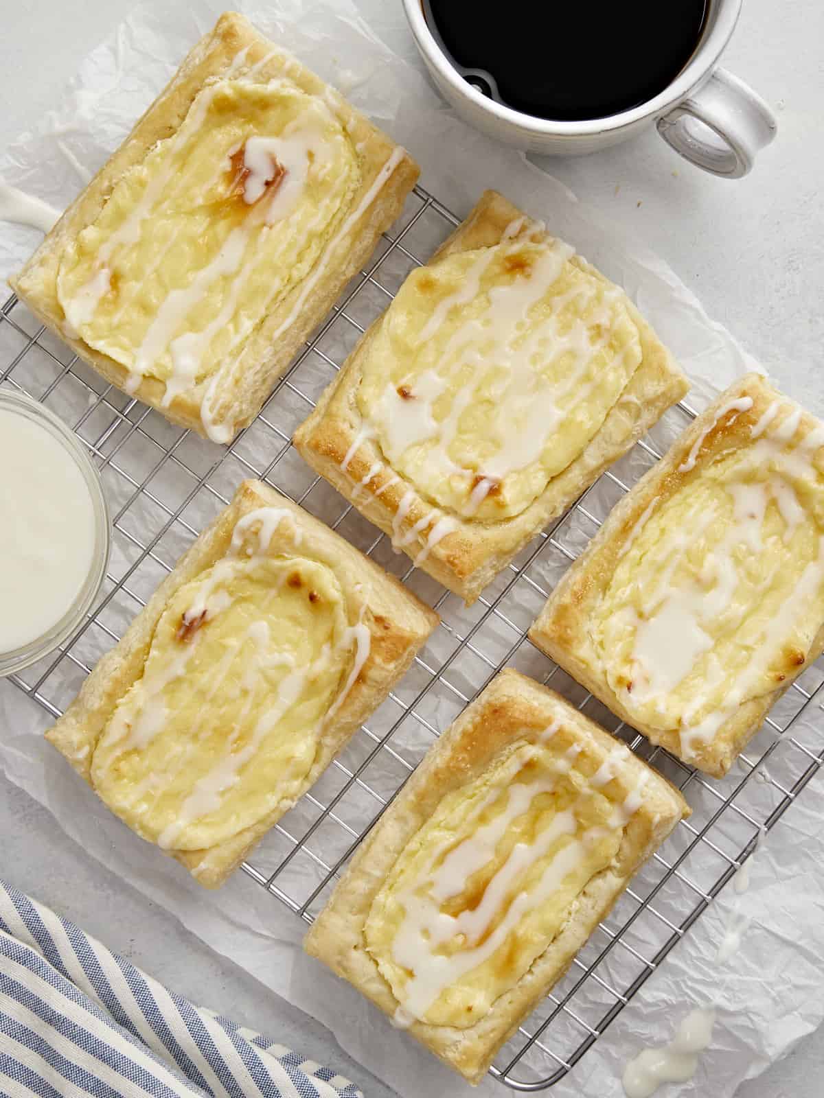 Overhead shot of cheese danish on a cooling rack with a small bowl of icing and a sup of coffee next to them.