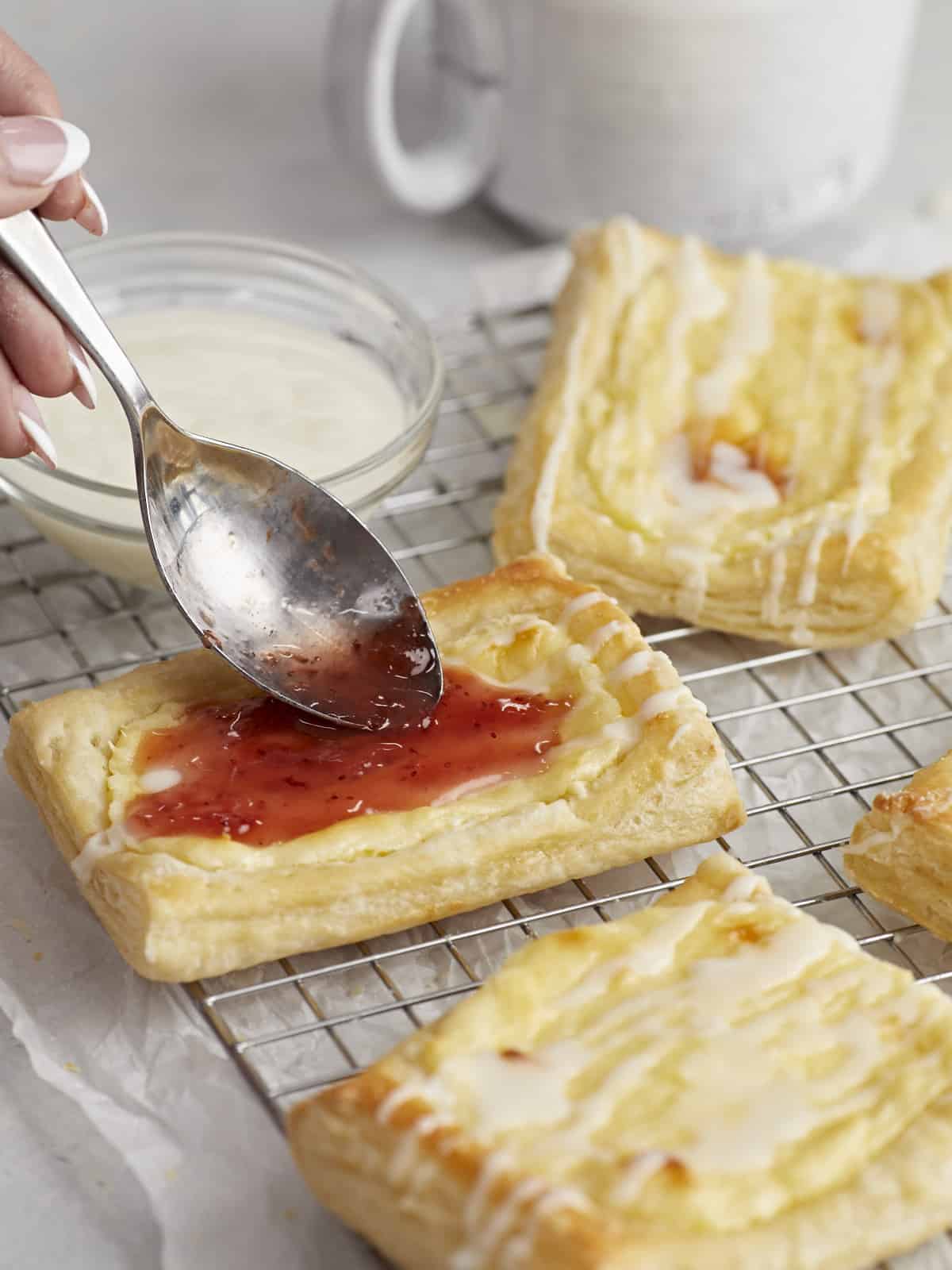 Side shot of cheese danish on a cooling rack with a small bowl of icing next to them wit hone danish being dressed with strawberry jam.
