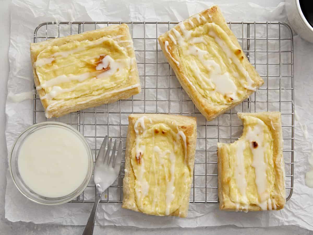 Overhead shot of cheese danish on a cooling rack with a small bowl of icing and a silver fork next to them.