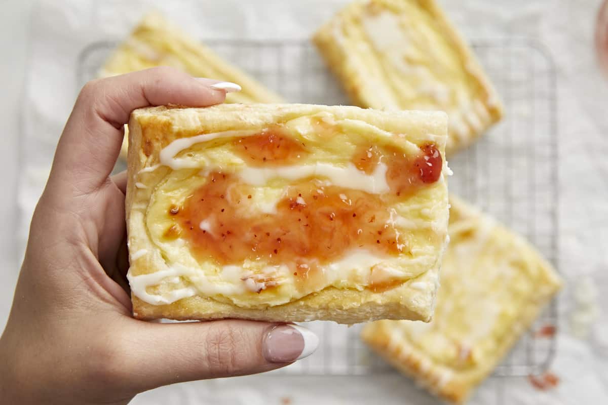 Overhead shot of a hand holding a cheese danish dressed with icing and strawberry jam in the foreground and three other cheese danish on a cooling rack in the background.