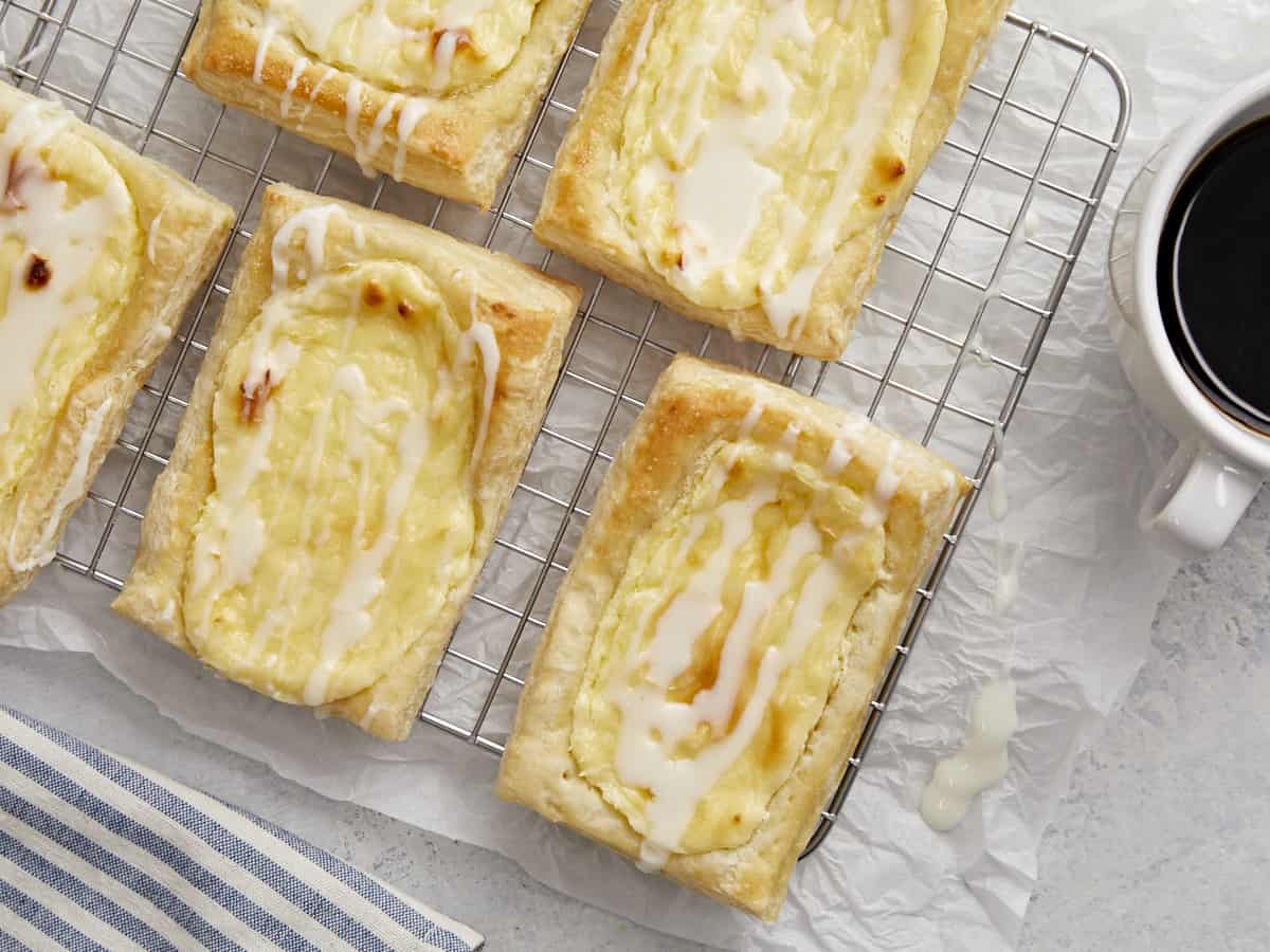 Overhead shot of cheese danish on a cooling rack with a small bowl of icing and a sup of coffee next to them.