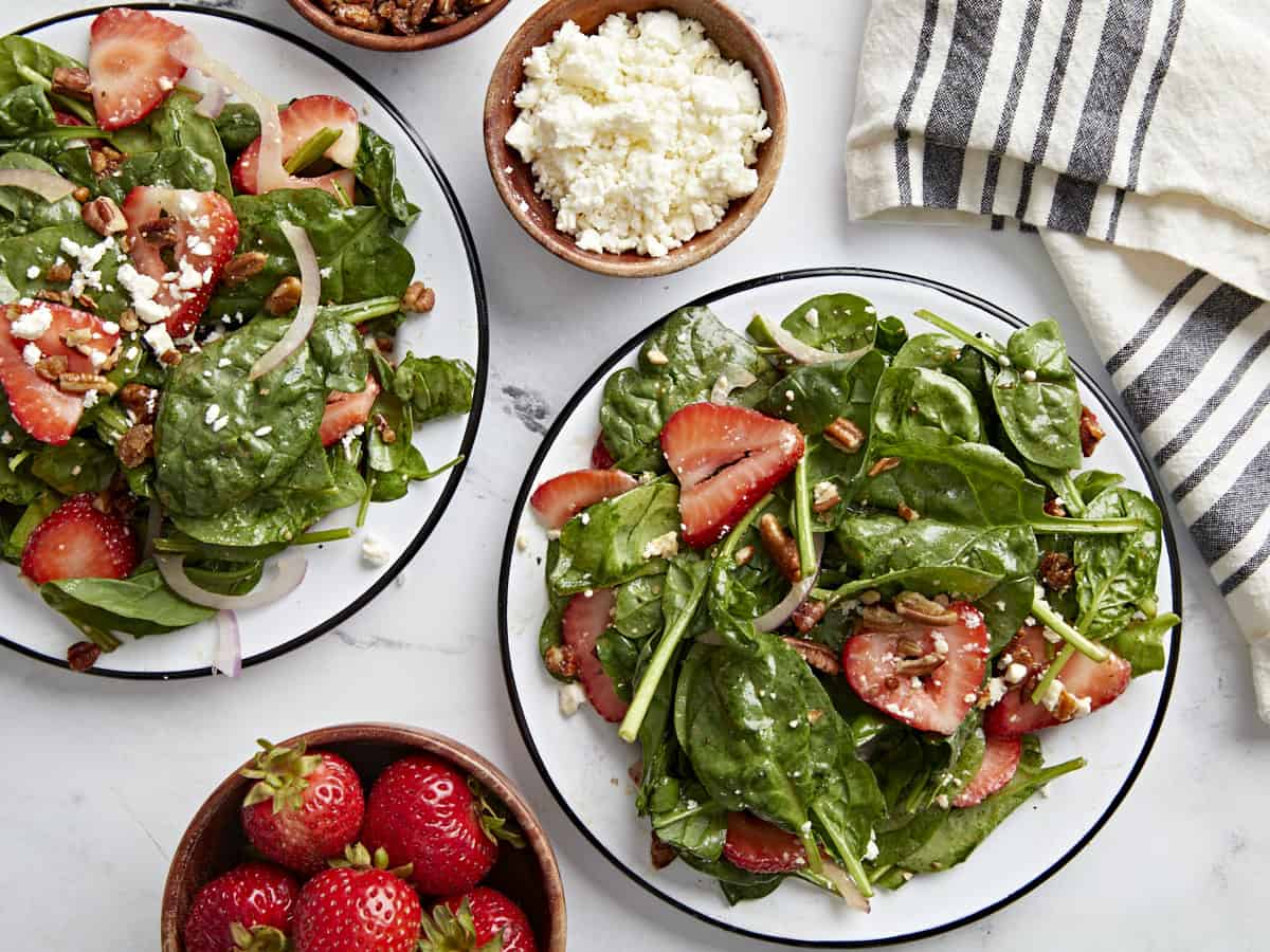 Overhead shot of two plates of strawberry spinach salad with a small bowl of strawberries and feta cheese on the side.