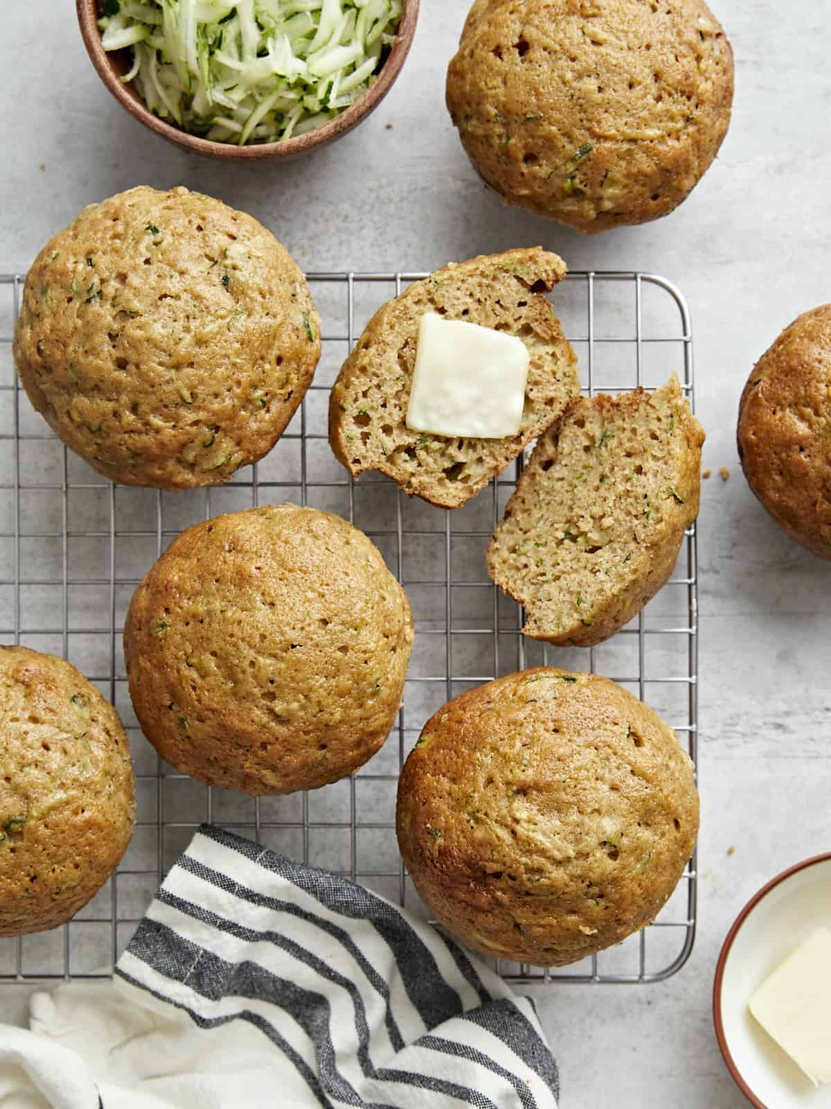 Overhead view of zucchini muffins on a wire rack with one cut open and butter spread in the middle.