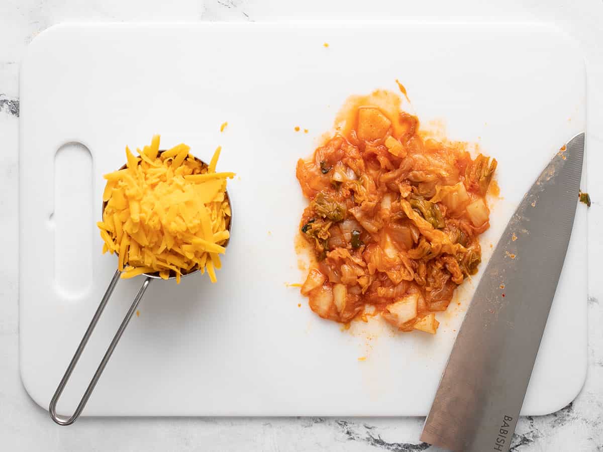 Chopped kimchi on a cutting board with shredded cheddar in a measuring cup on the side.