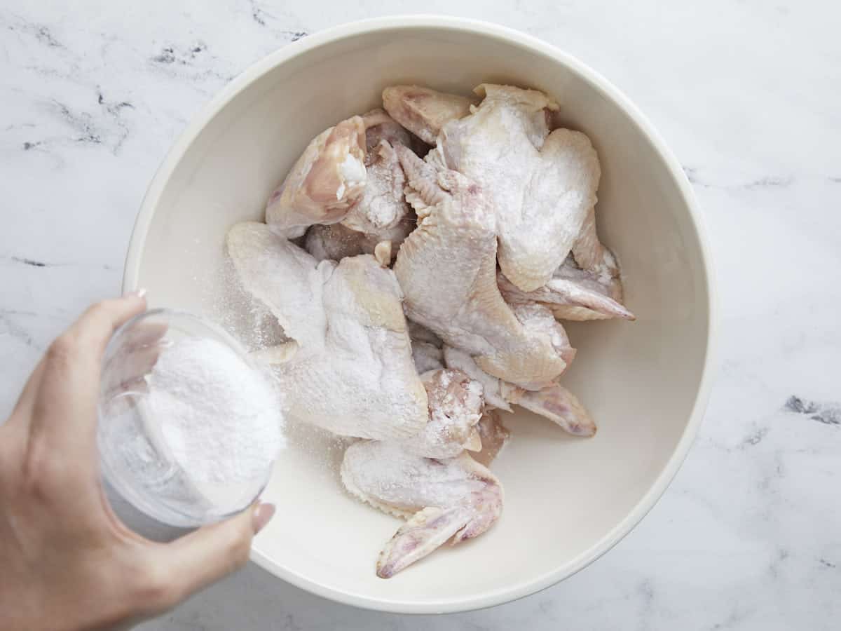 Overhead shot of raw chicken wings in a white bowl being sprinkled with baking powder and salt.