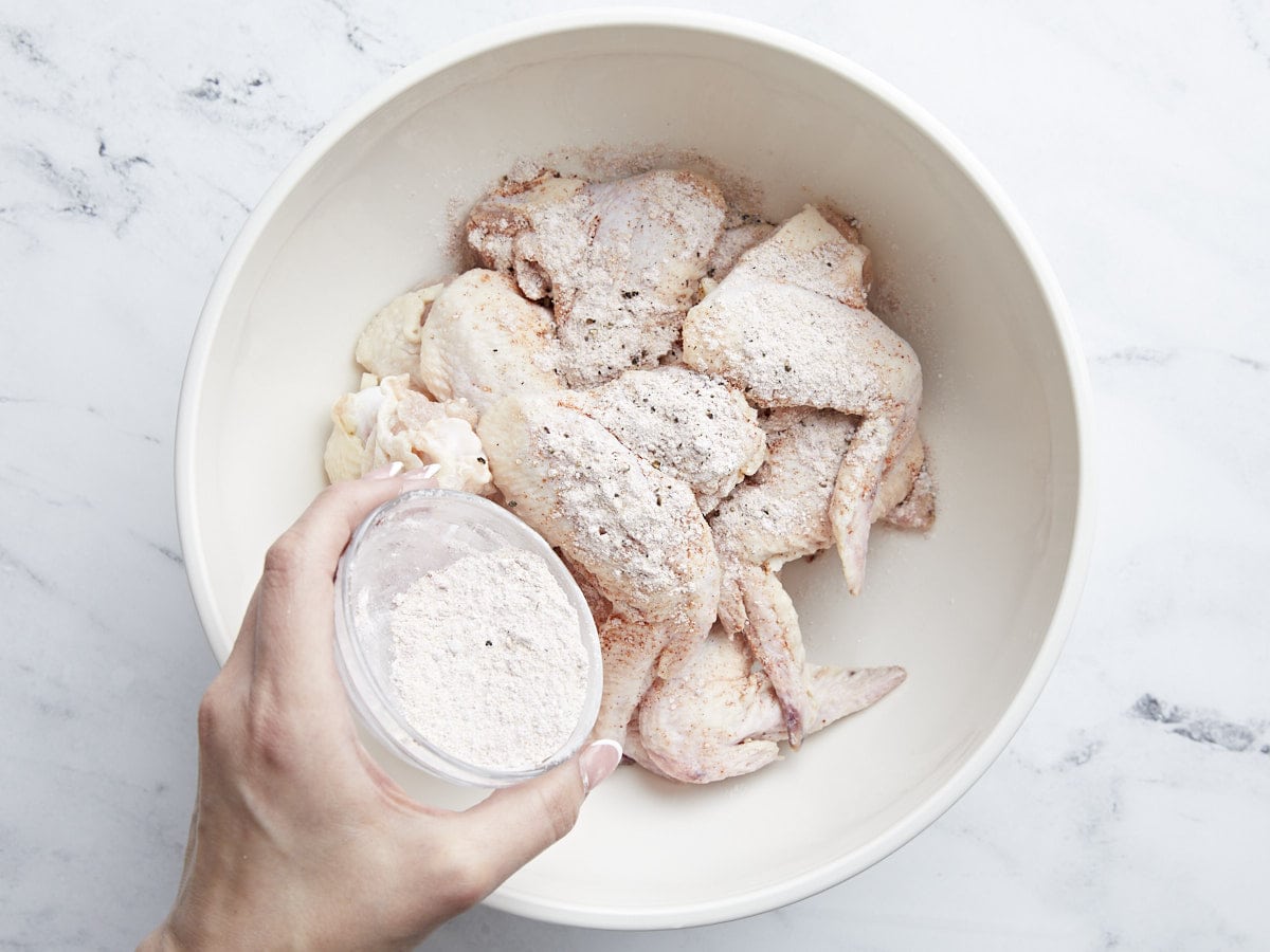 Overhead shot of raw chicken wings in a white bowl being sprinkled with cornstarch and seasonings.