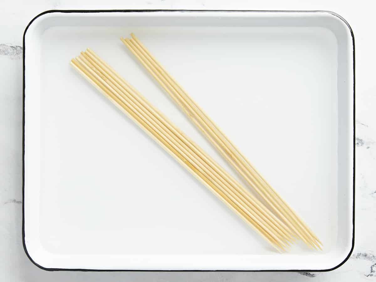 Overhead shot of wood skewers soaking in water in a white sheet pan.