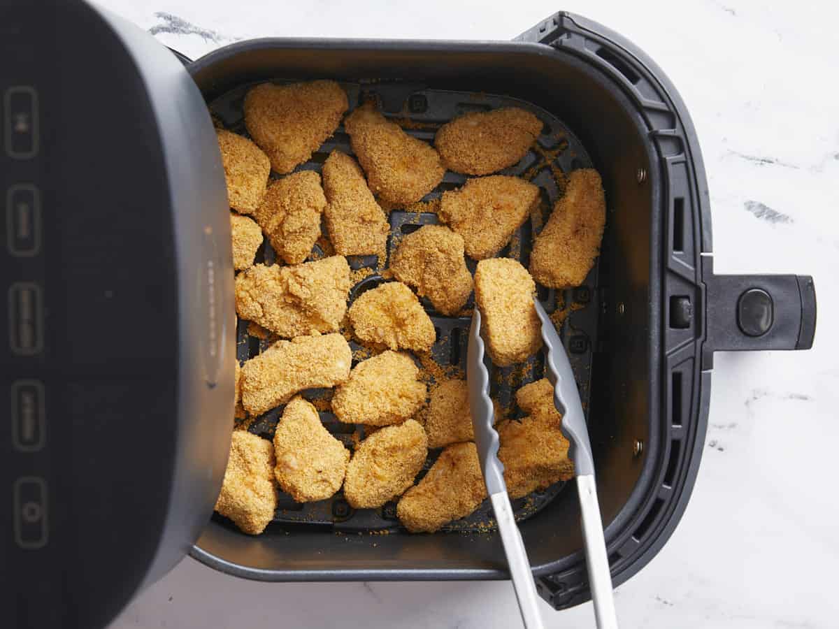 Cooked chicken nuggets in the air fryer basket with one being lifted with tongs.