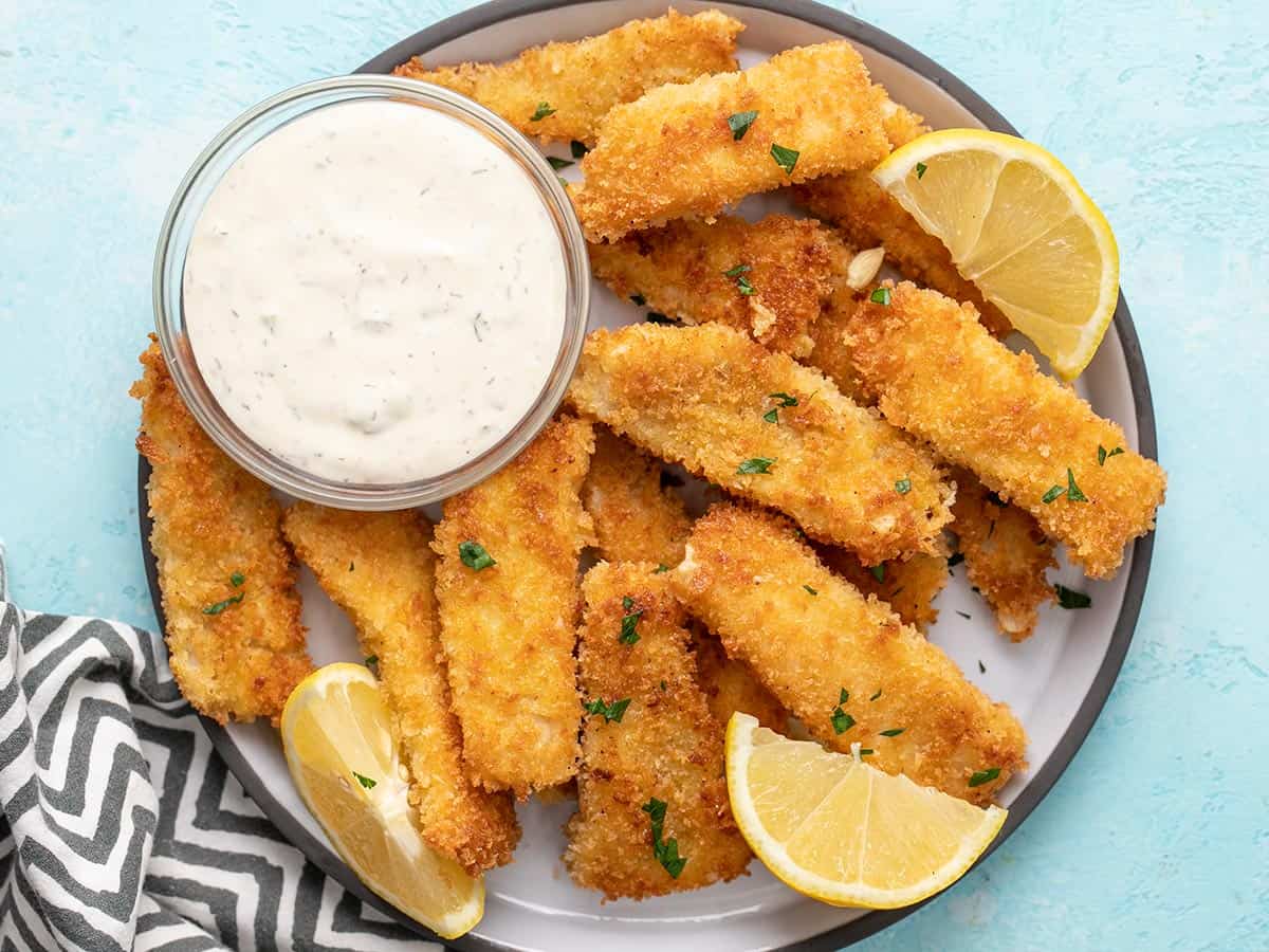 Overhead view of a plate full of fish sticks with tartar sauce and lemon wedges.