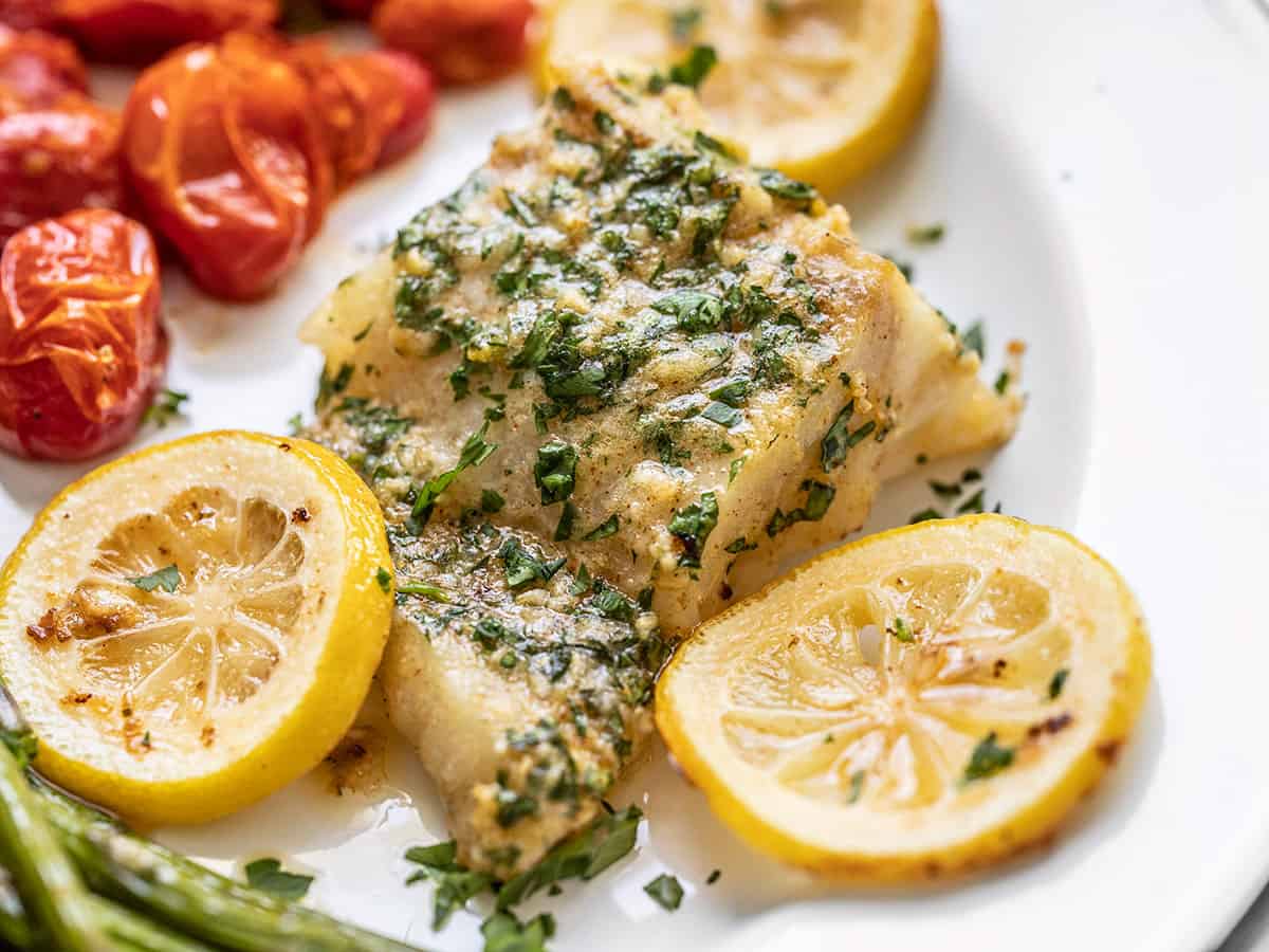 Close up side view of a piece of garlic butter baked cod on a serving plate with asparagus and tomatoes.