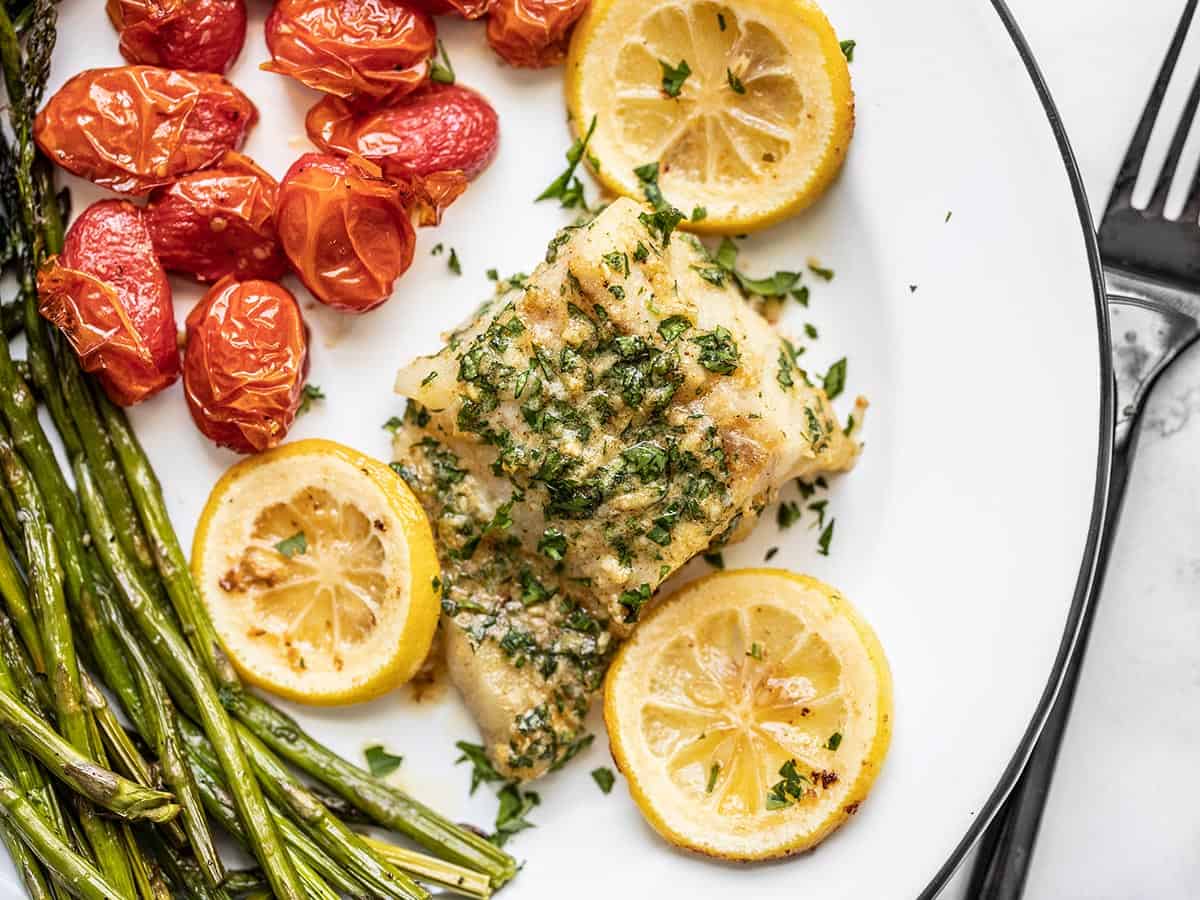 Overhead view of a piece of garlic butter baked cod on a plate with roasted asparagus and tomatoes.