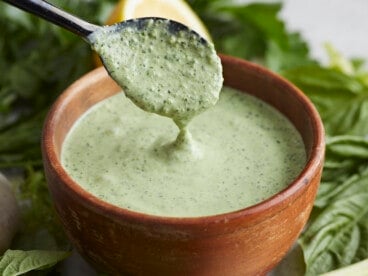 Side shot of Green Goddess Dressing in a wood bowl with a black spoon drizzling some in.