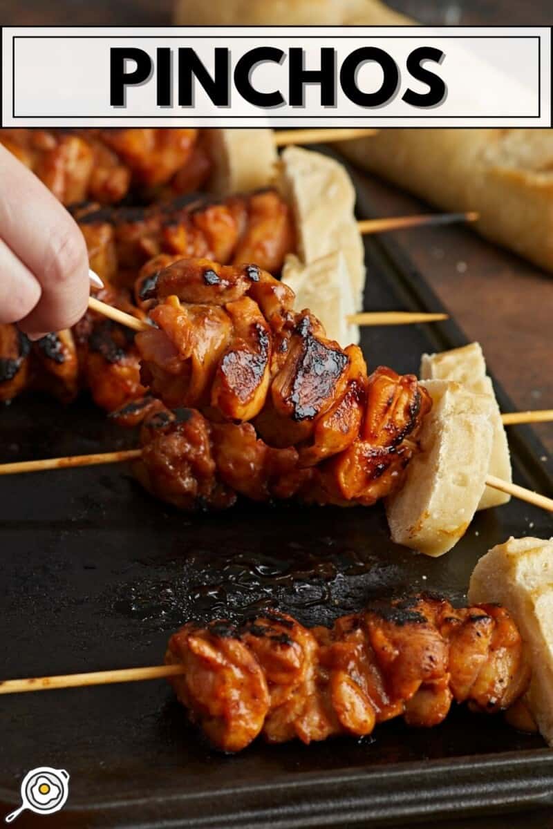 Side shot of hand lifting Pinchos de Pollo on a black grill plate.