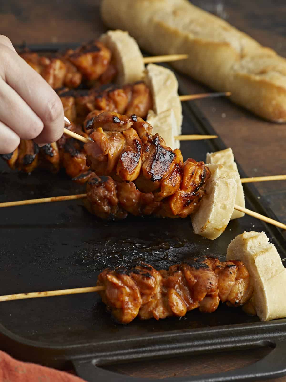 Side shot of hand lifting Pinchos de Pollo on a black grill plate with a loaf of bread in the back ground.