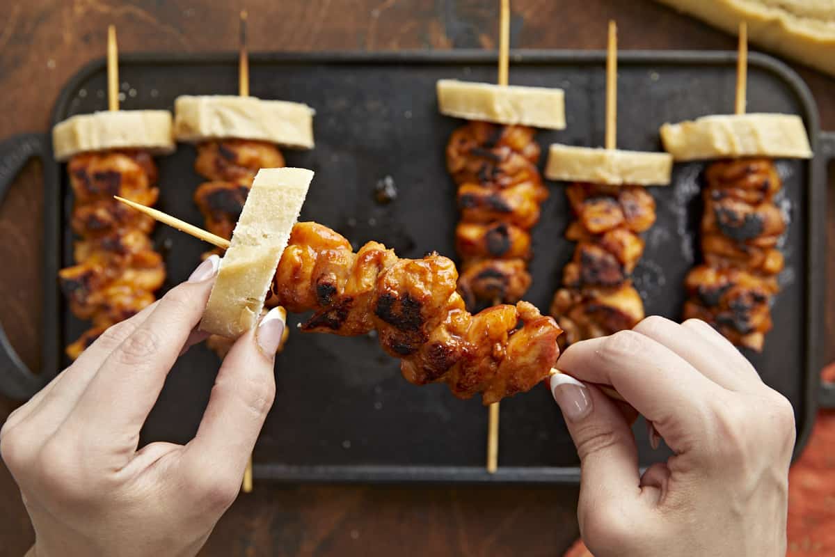 Over head shot of hand lifting Pinchos De Side shot of hand lifting Pinchos de Pollo on a black grill plate.