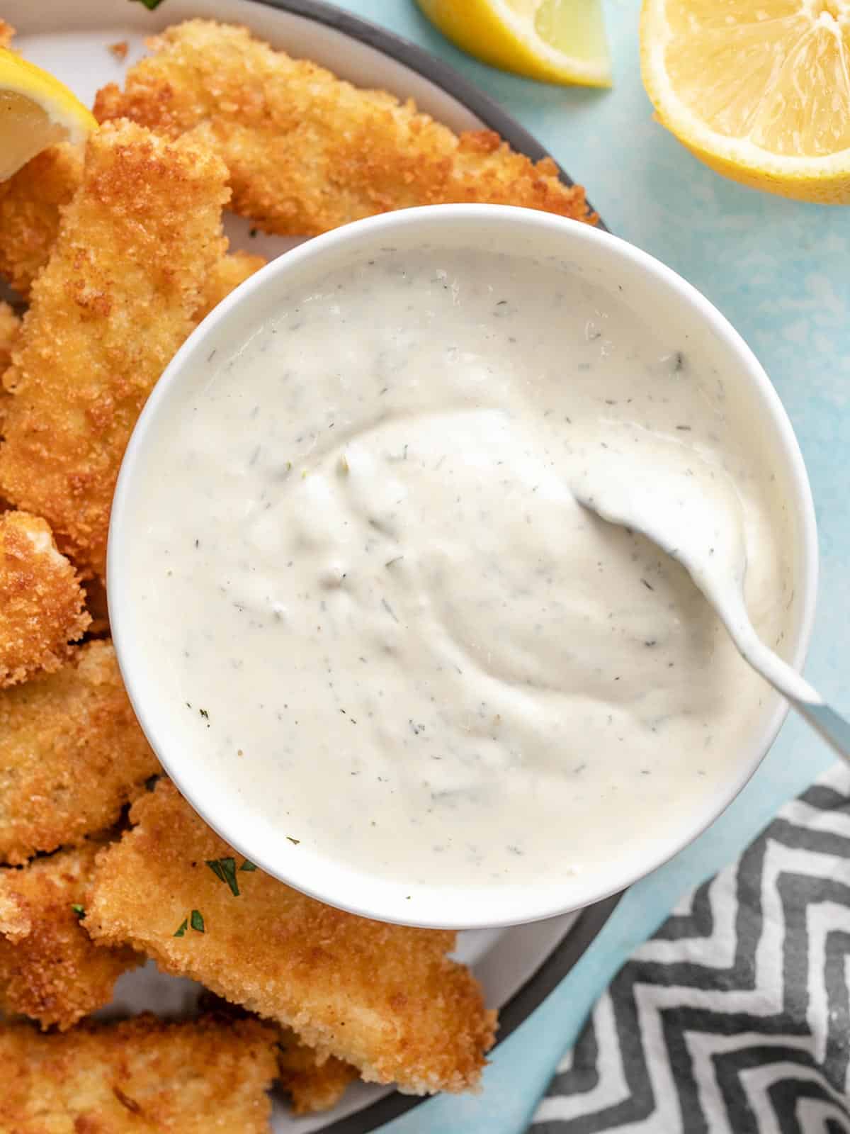 Overhead view of a bowl of tartar sauce on a plate of fish sticks, being stirred with a spoon.