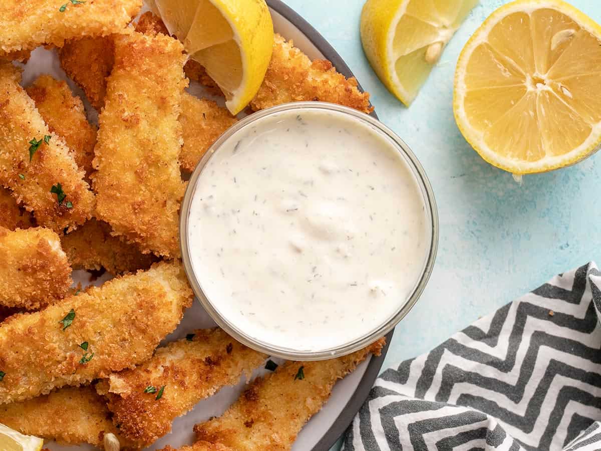 A bowl of homemade tartar sauce on a plate of fish sticks with lemons on the side.