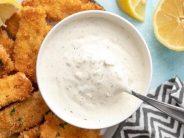 Overhead view of a bowl of tartar sauce with a spoon, on a plate of fish sticks.