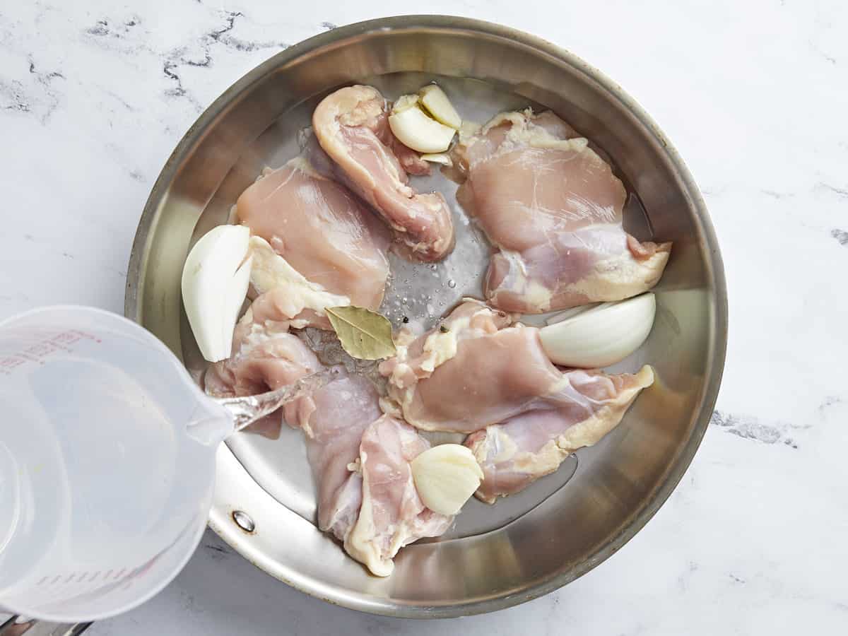 Chicken thighs in a pan with onion and garlic, water being poured in.