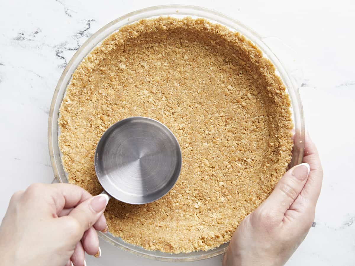 Cookie crumbs being pressed into a pie plate with the back of a measuring spoon.
