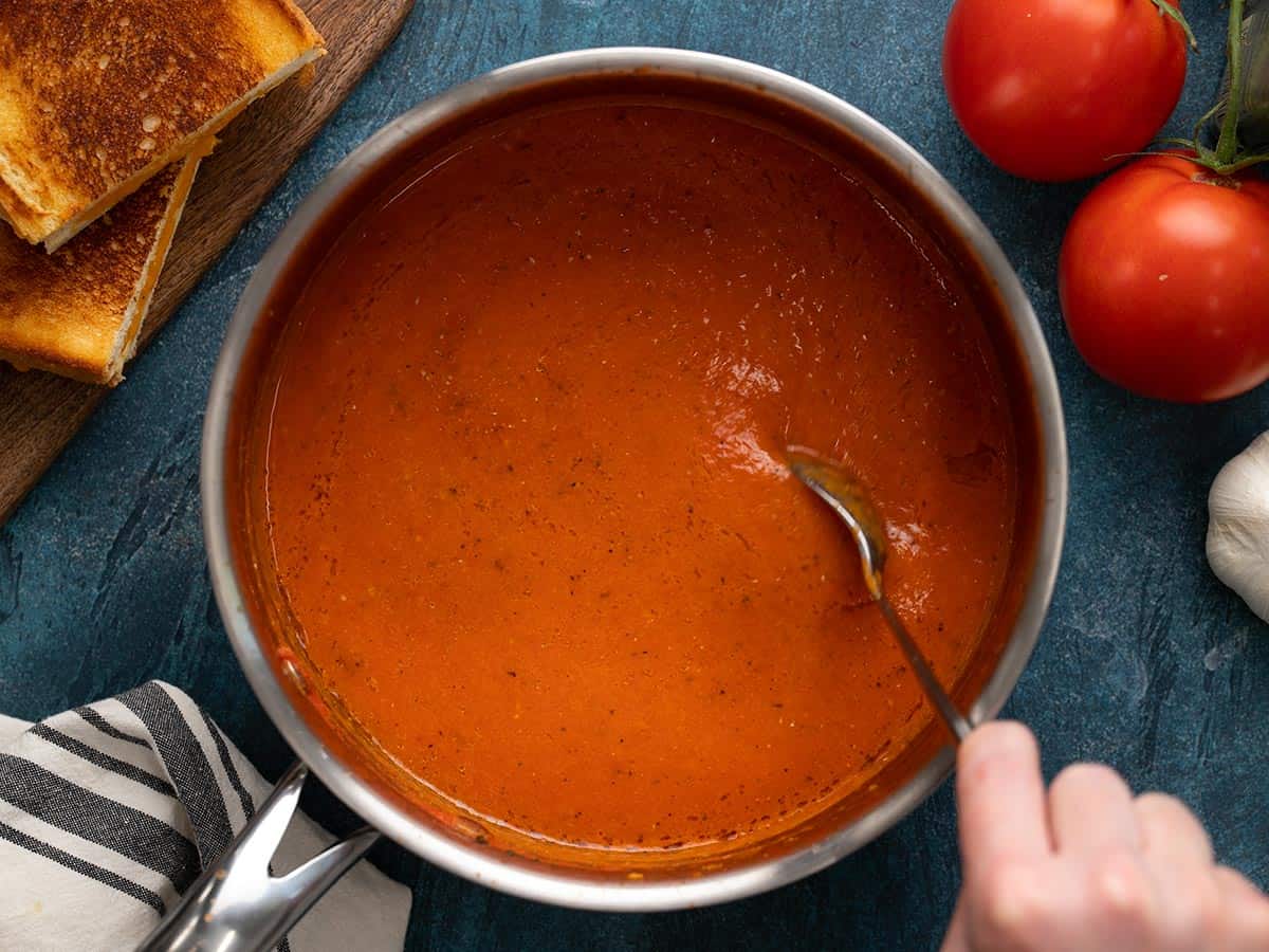 A pot of roasted tomato soup being stirred with a spoon.