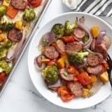 A serving of Sheet Pan Chicken Sausage and roasted vegetables on a white plate next to a sheet pan.