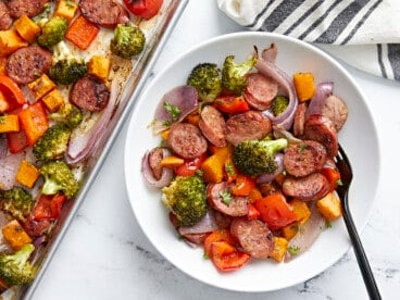 A serving of Sheet Pan Chicken Sausage and roasted vegetables on a white plate next to a sheet pan.