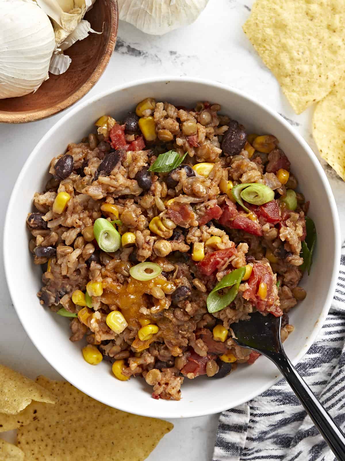 Overhead view of a white bowl full of southwest lentils and rice with tortilla chips, a napkin, and black fork on the side.