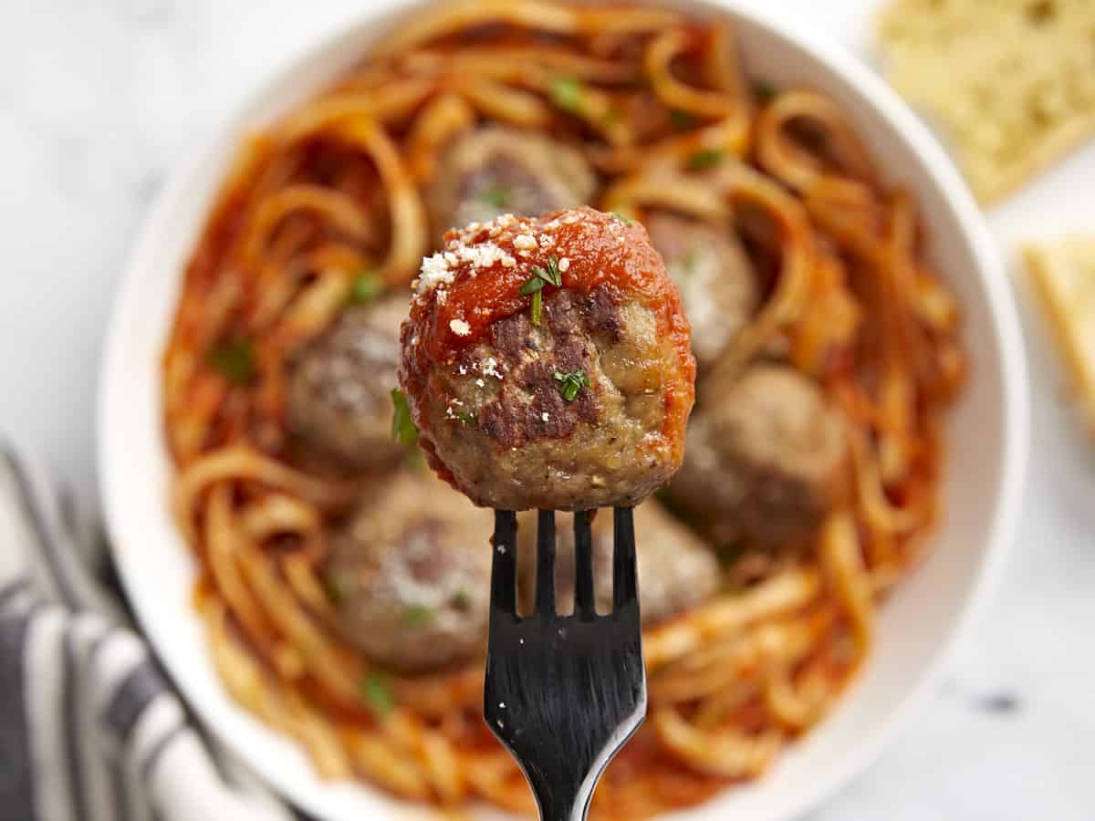 Close up of a turkey meatball on a fork with a bowl of pasta in the background. 