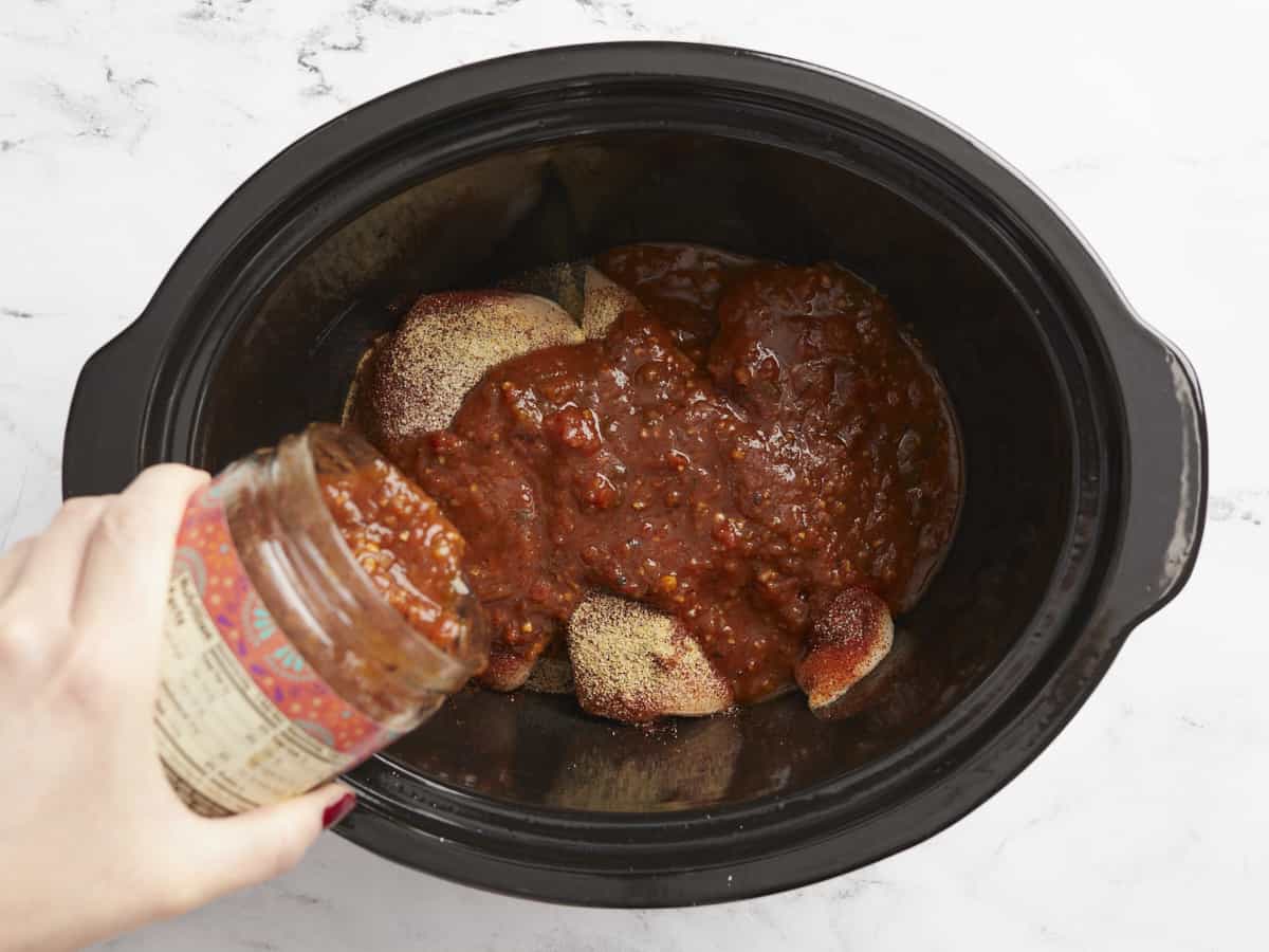 A jar of salsa being poured over top of chicken in the slow cooker.