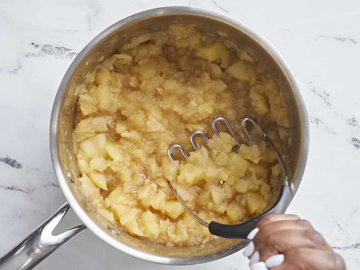 Overhead view of cooked apples in a saucepan being mashed with a potato masher.
