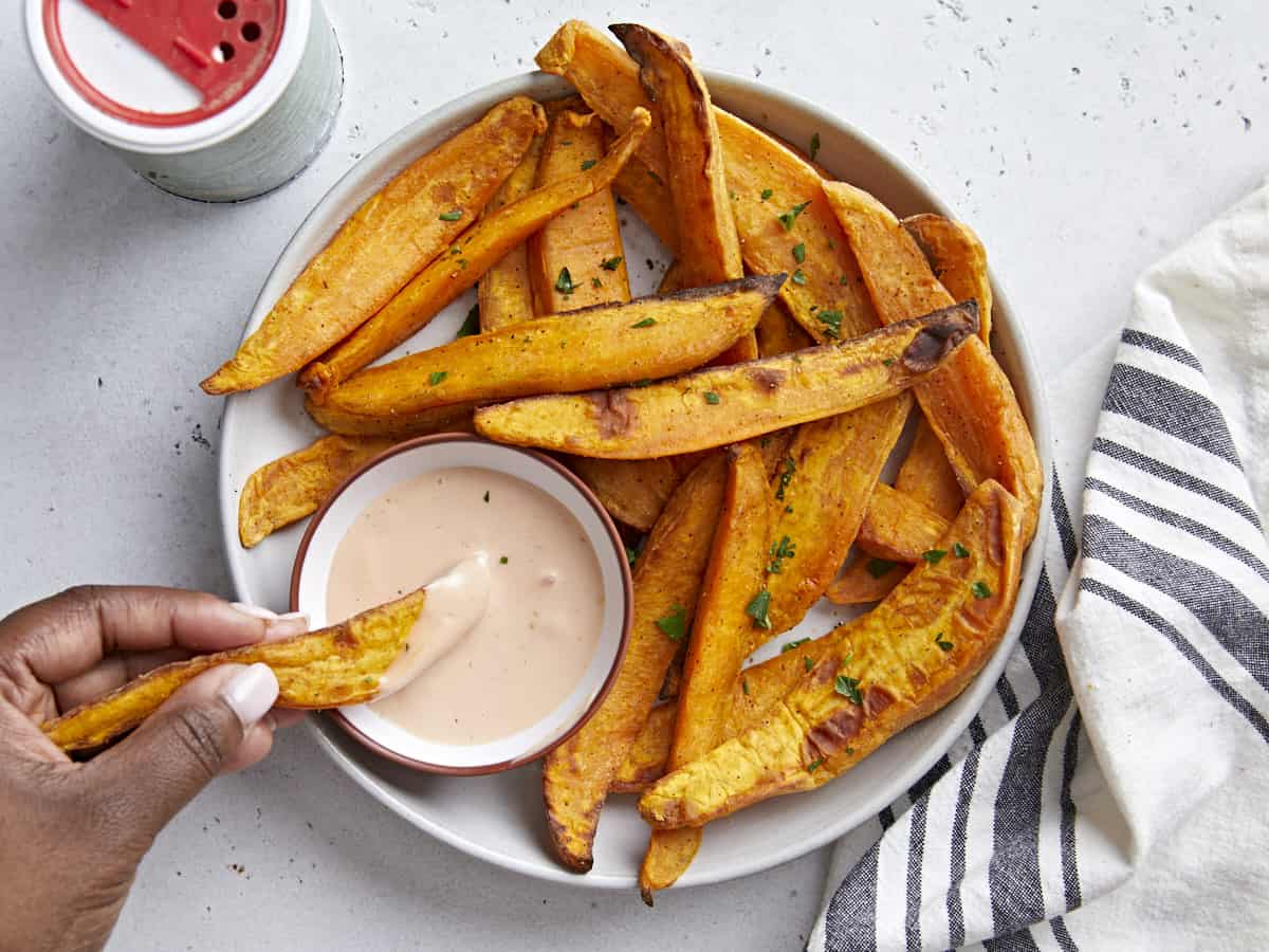 Overhead view of sweet potato fries on a serving plate with one fry being dipped in a side of mayo ketchup.