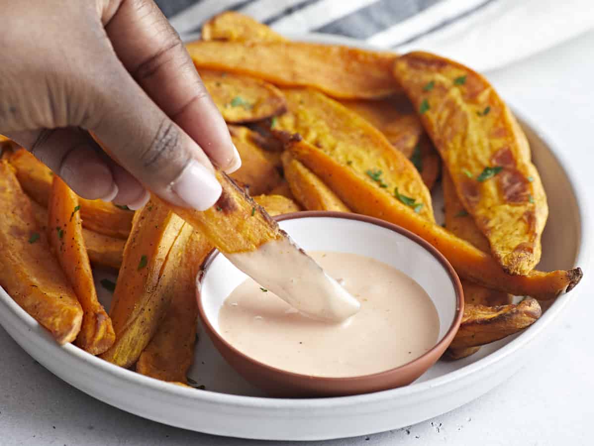 Side view of sweet potato fries on a serving plate with one fry being dipped in a side of mayo ketchup.