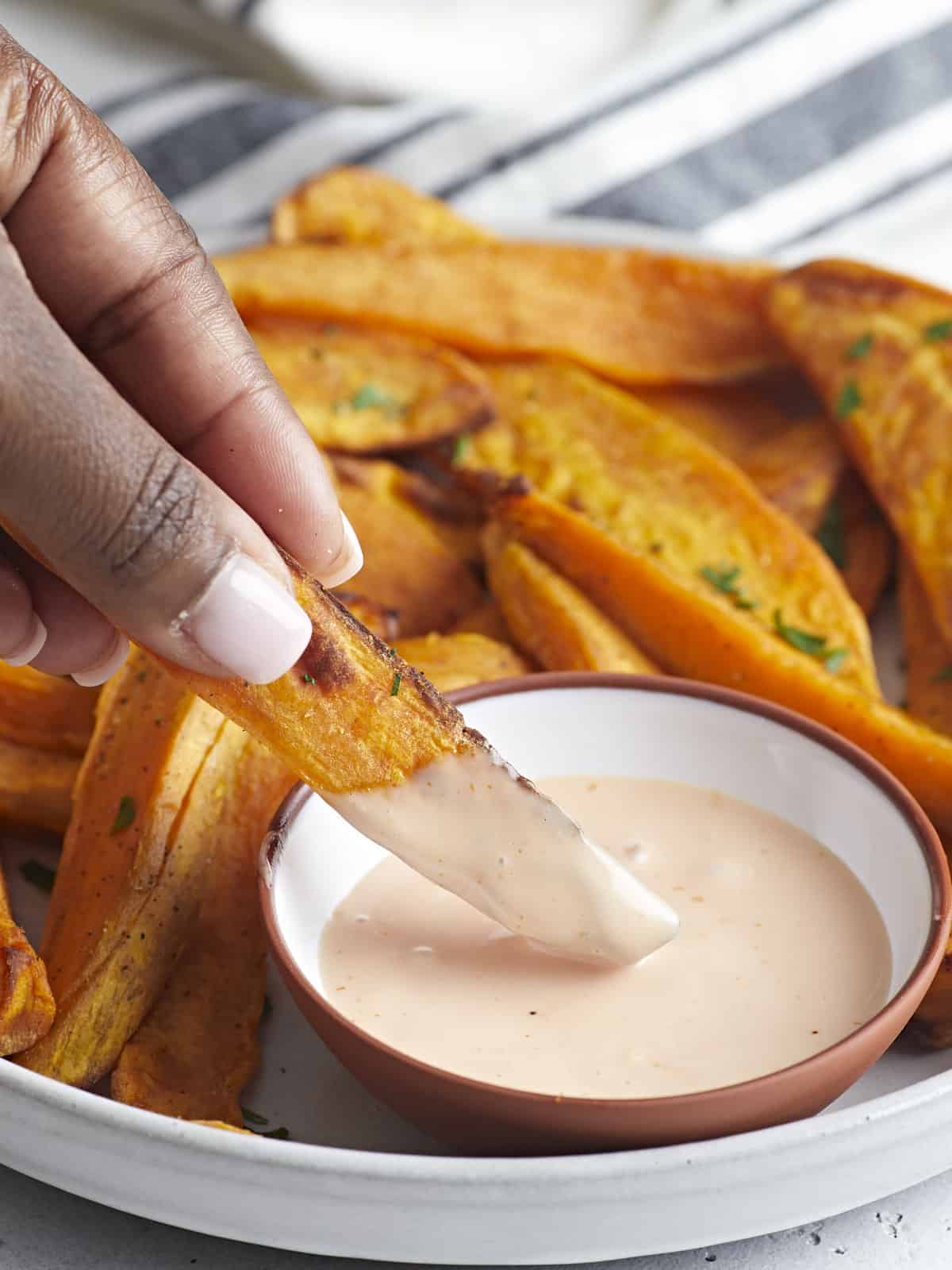 Side view of sweet potato fries on a serving plate with one fry being dipped in a side of mayo ketchup.