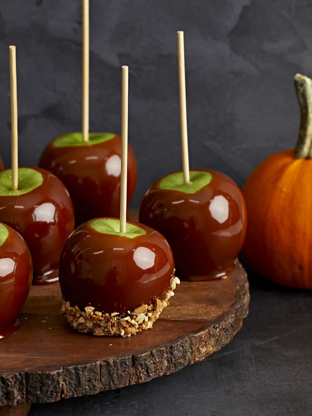 Caramel apples on a wooden platter with a pumpkin on the side.