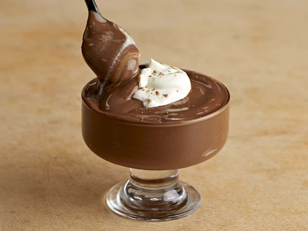 Side view of a spoon dipping into a bowl of chocolate pudding with whipped cream on top.