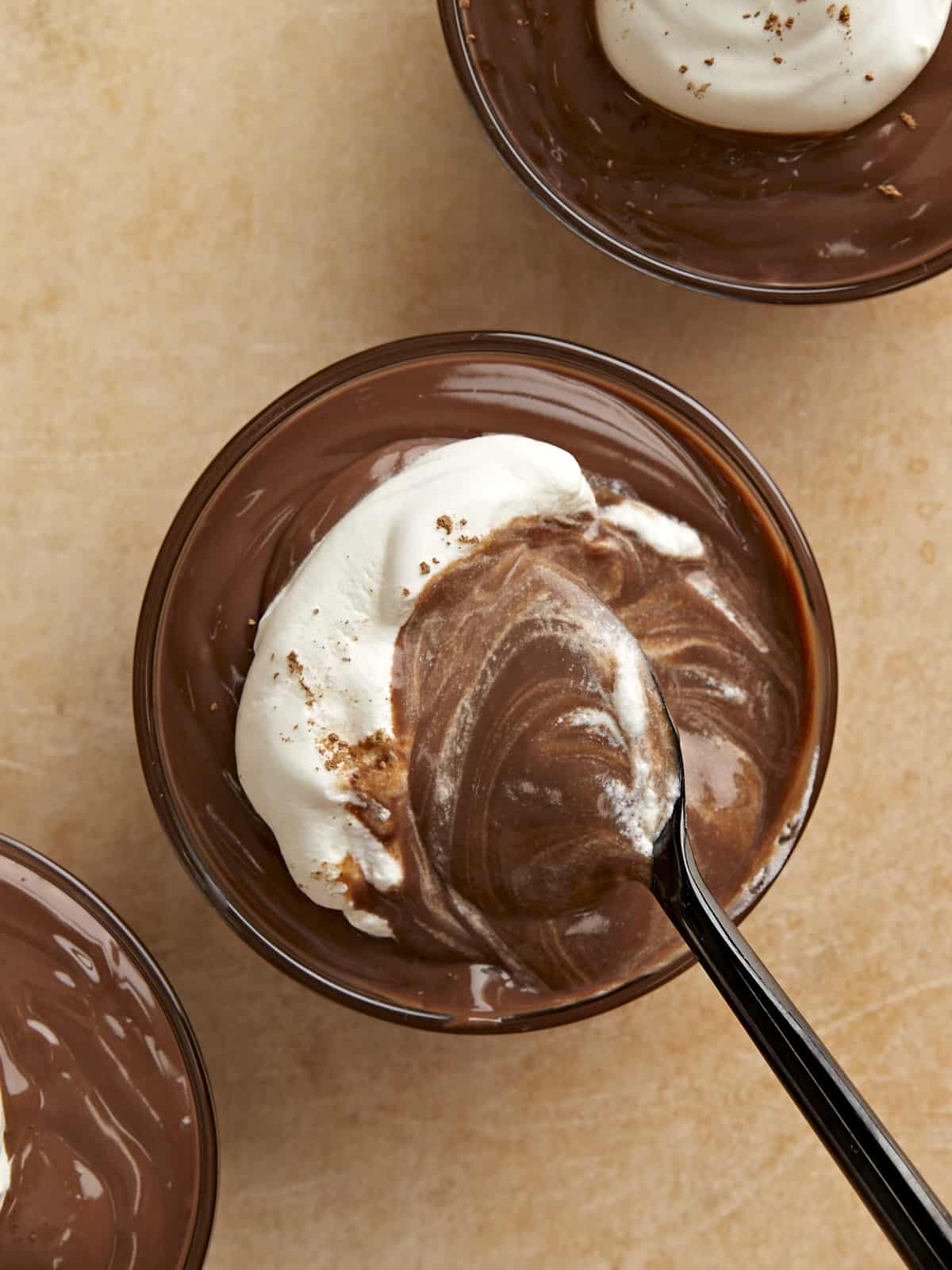 Overhead view of a bowl of chocolate pudding with whipped cream on top and a spoon stirring.