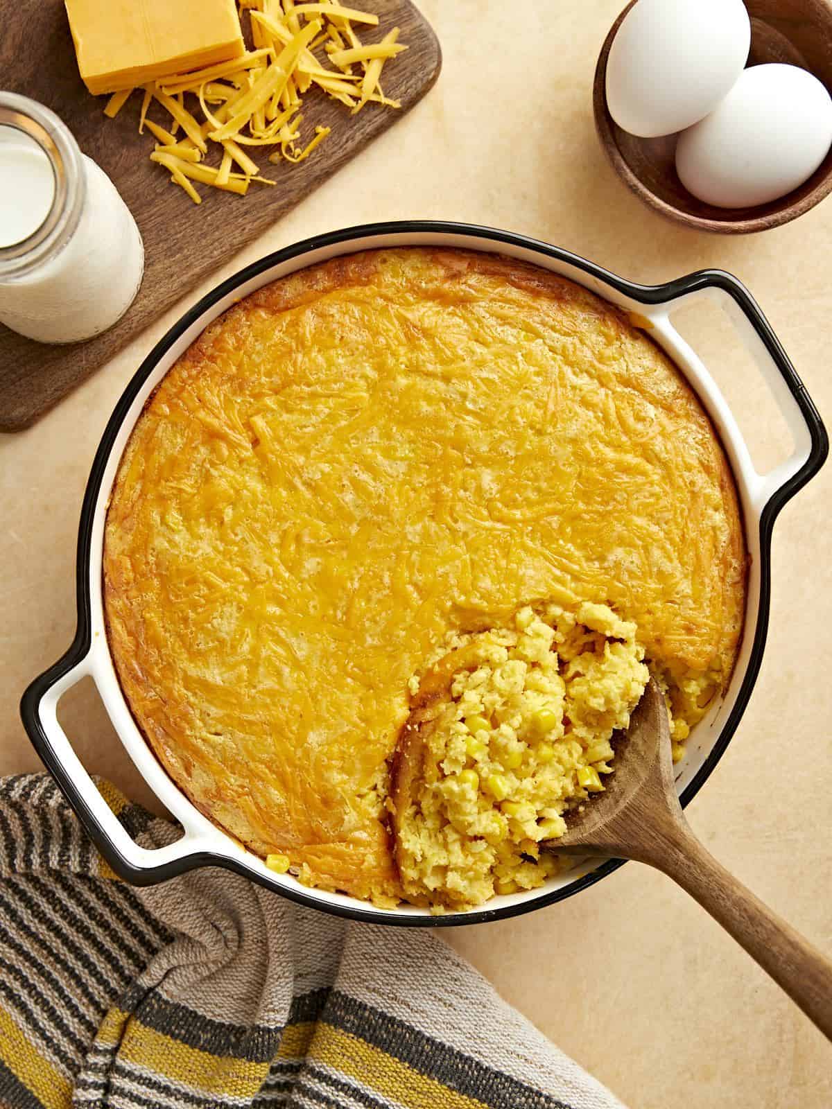 Overhead view of corn pudding being scooped out of the casserole dish with ingredients on the sides.