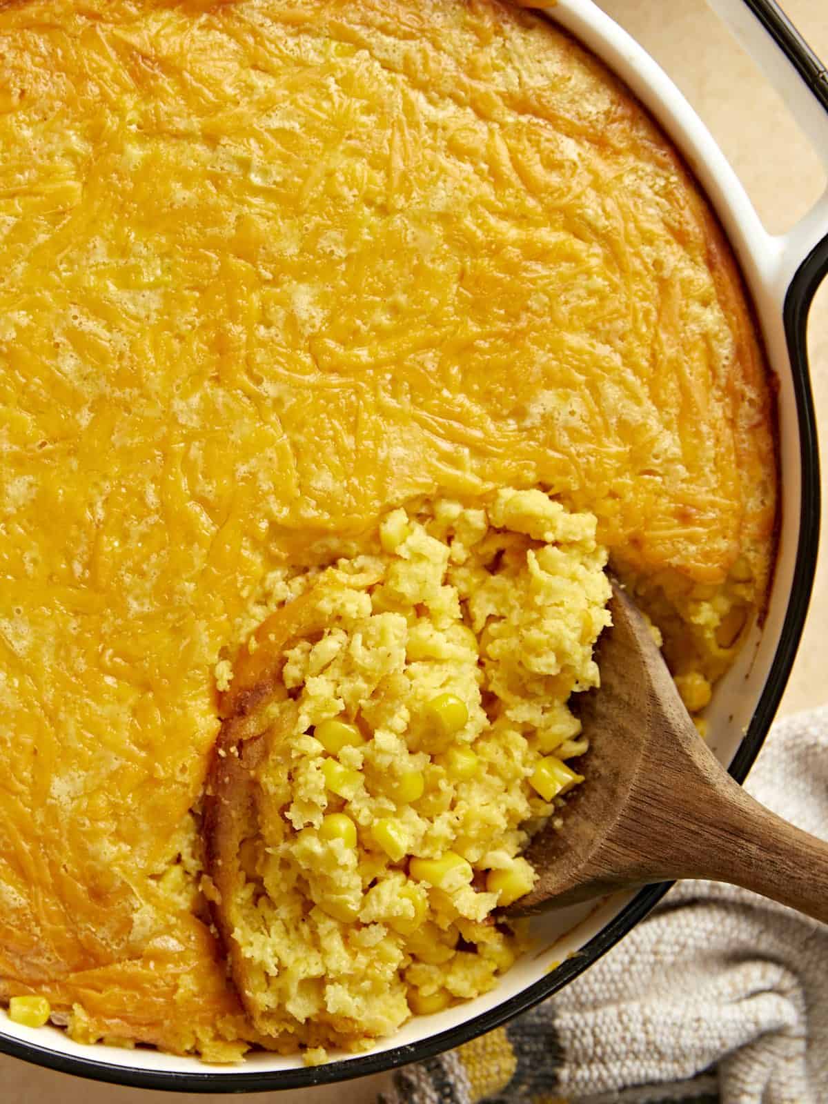 Close up overhead view of corn pudding in the casserole dish with a wooden spoon scooping some out.